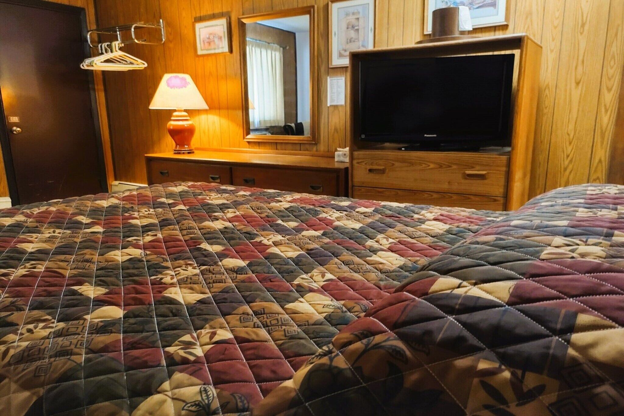 Hotellihuone, jossa on suuri sänky, televisio, puinen lipasto, peili ja lamppu.