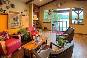 A cozy living room with a red leather sofa, a wooden coffee table, and a large window overlooking a scenic view. - Waterfront Gem on the River! Hot Tub & Boat Dock! (Priest River)
