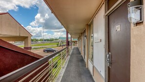Un pasillo con una galería techada, una puerta con el número 221, y vista a un estacionamiento y vegetación.