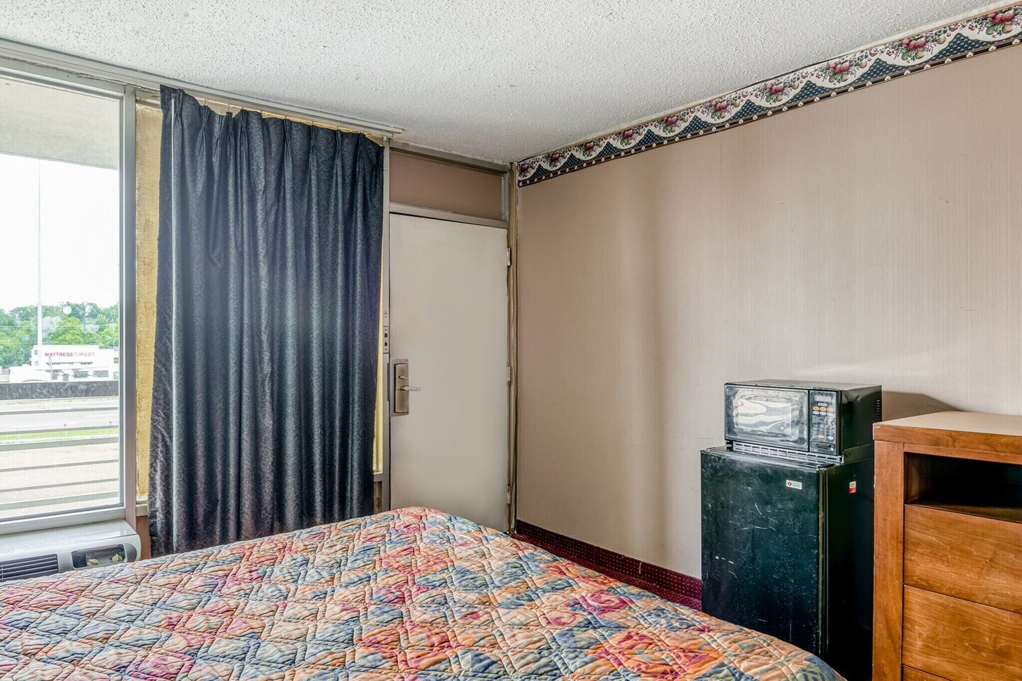Une chambre d’hôtel avec un lit, un petit réfrigérateur, une commode en bois, une fenêtre avec des rideaux et une porte.