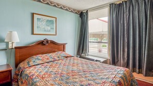 Habitación de hotel con cama, mesita de noche, lámpara, ventana con cortinas y un cuadro en la pared.