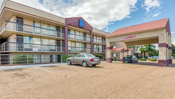Un hotel de varios pisos con área de recepción, estacionamiento y un automóvil estacionado al frente.