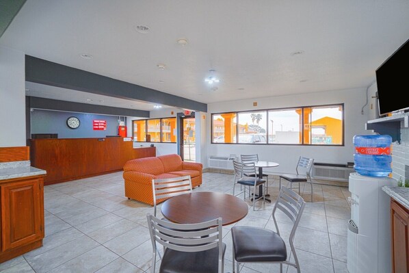 A hotel lobby with a reception desk, a sofa, dining tables, chairs, and a large water dispenser.