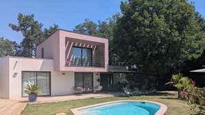 Una casa moderna de dos pisos con piscina, mobiliario exterior y un gran árbol al fondo.