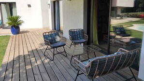 Terraza de madera con sillas de mimbre y una mesa de vidrio, con vista a una piscina.