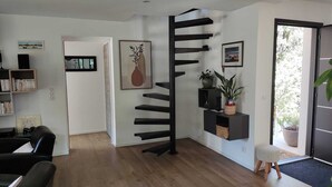 Una sala de estar moderna con piso de madera, una escalera espiral negra, una estantería y una puerta que da a un balcón con plantas en macetas.