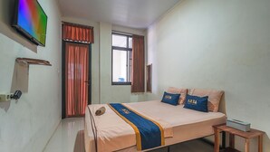 A bedroom with a bed, pillows, a bedside table, a TV, curtains, a window, and a wall-mounted shelf. - Hotel Danau Tjokro 3 By ARBA (Cisarua)