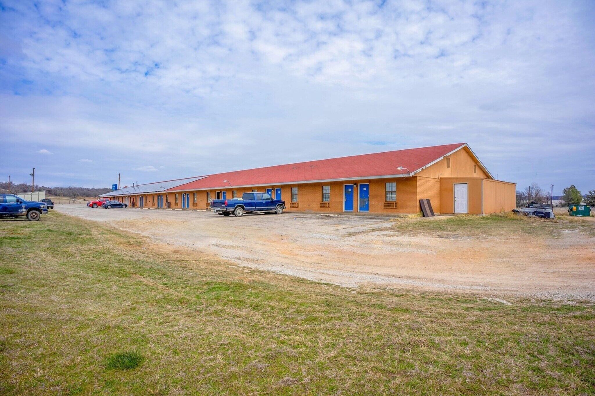 Un motel con techo rojo, varias puertas azules y un estacionamiento de grava.