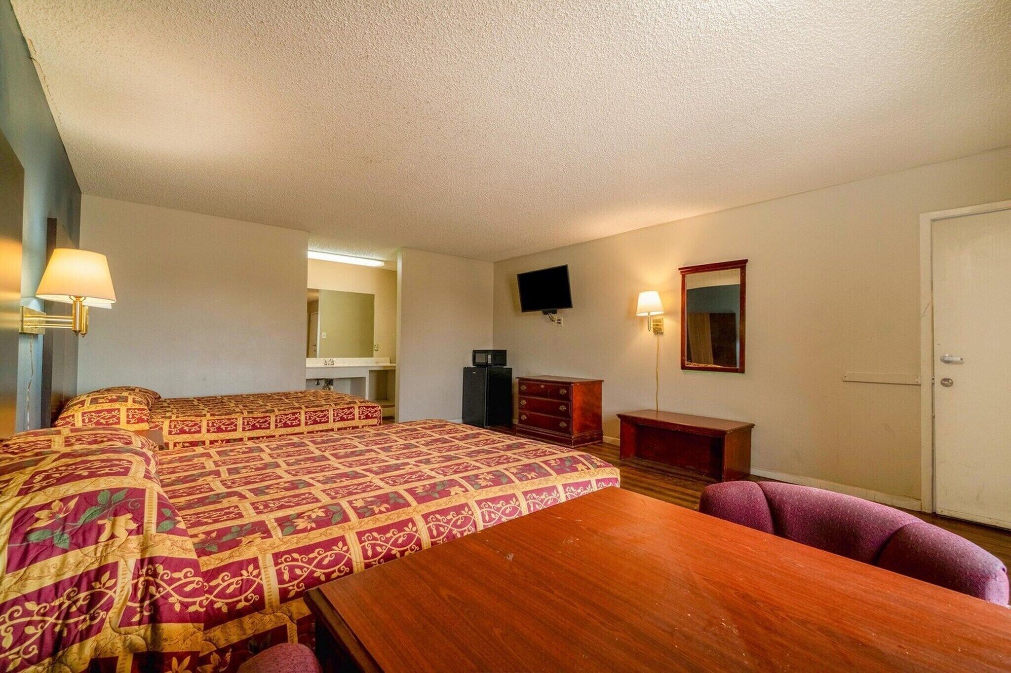A hotel room with a large bed, a wooden table, a desk, a television, a mirror, and a door.