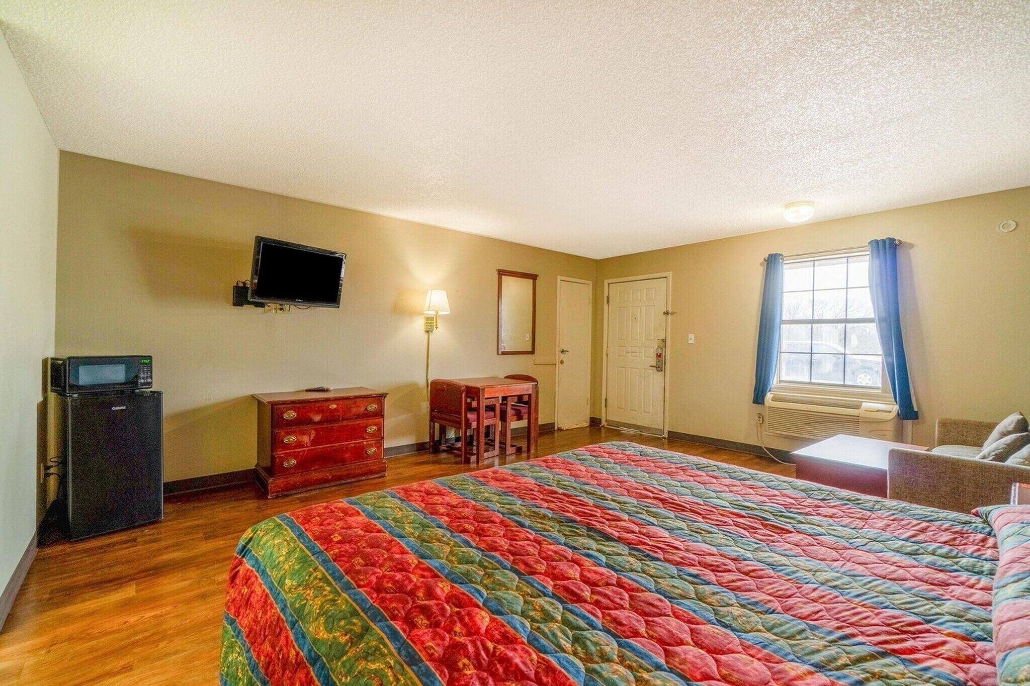 Un dormitorio con una cama grande, un televisor en la pared, un aparador, un escritorio y una ventana con cortinas.