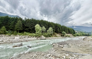 A river with rocks and trees, a mountain in the background, and cloudy sky. - Aranya Retreat Himachal (Mananli)