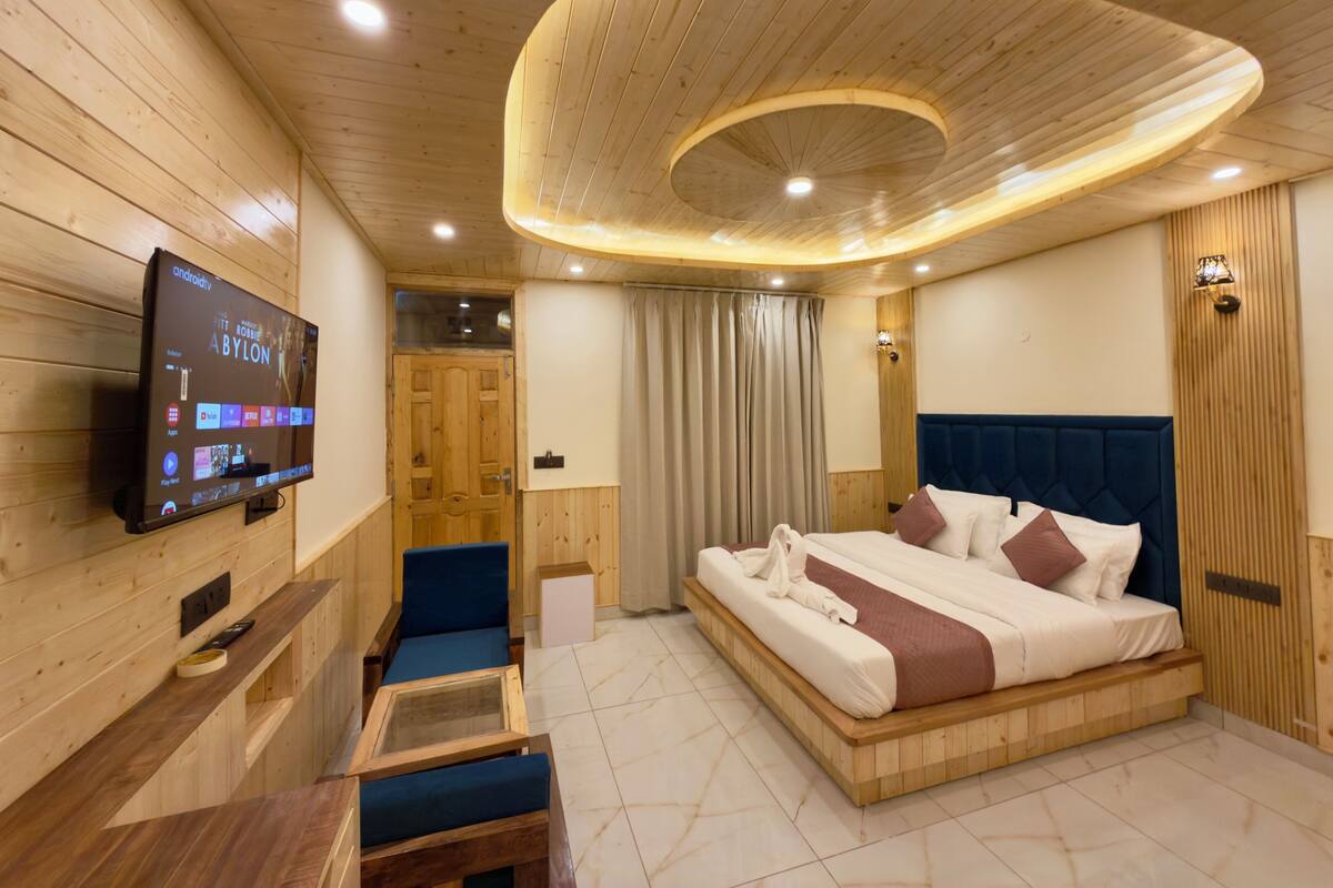 A modern hotel room with a wooden ceiling, a large bed, a television on the wall, and a small seating area.