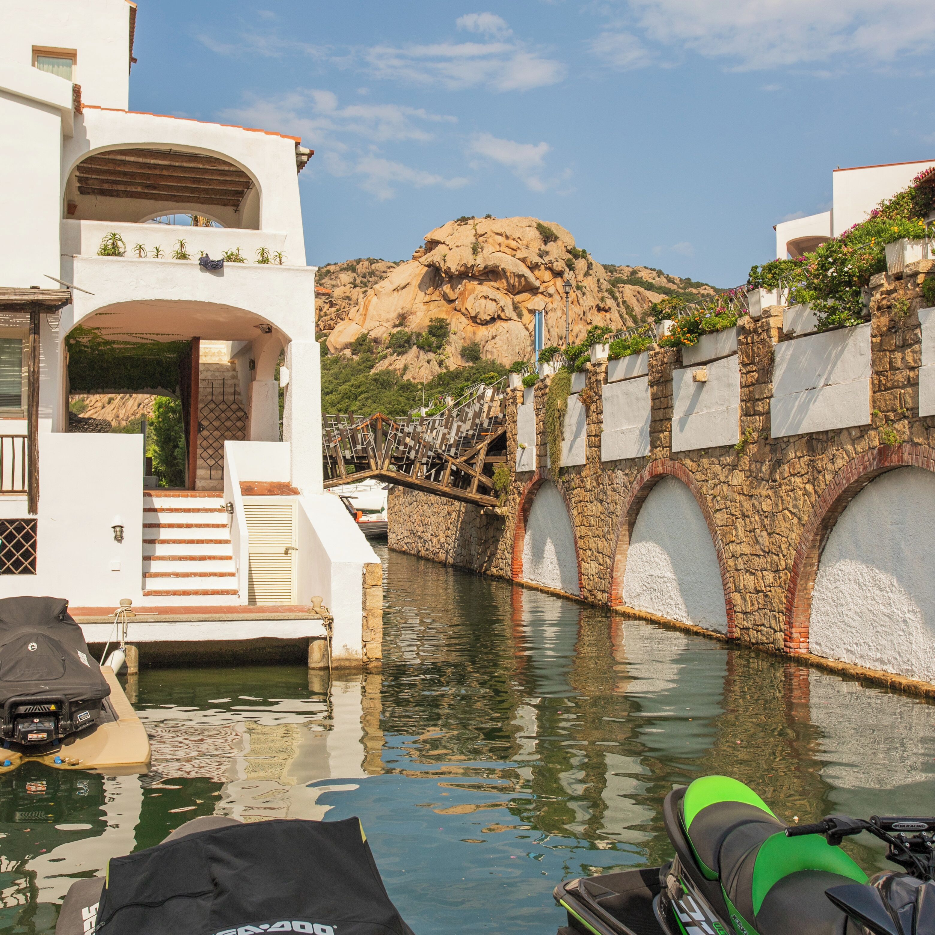 Un canale con un ponte di pietra, edifici bianchi con balconi e una montagna sullo sfondo.
