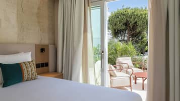 A hotel room with a bed, a wooden headboard, a nightstand, a window with curtains, and an outdoor seating area with chairs and a table.