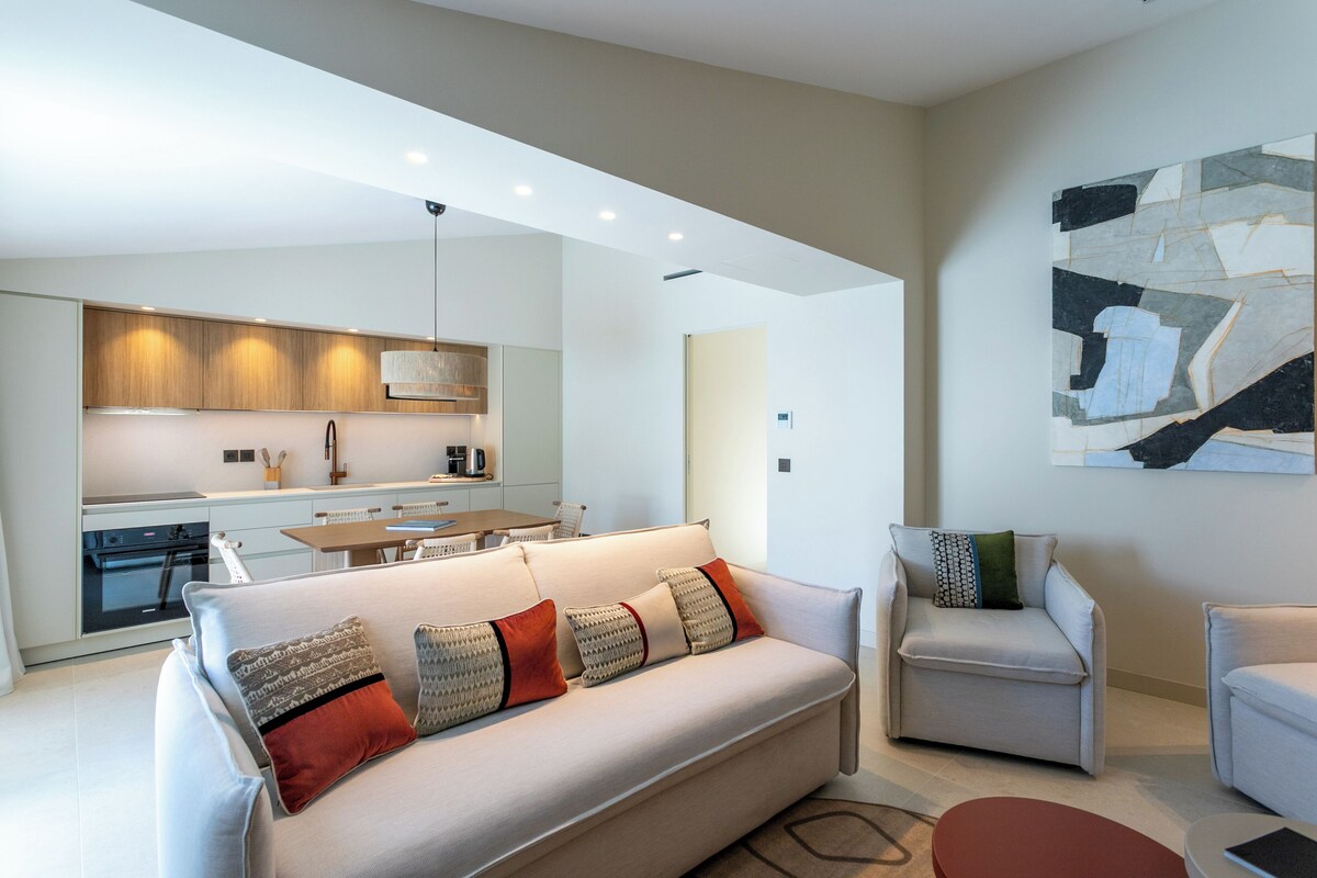 A modern living room with a sofa, armchairs, and a dining area with a kitchen in the background.