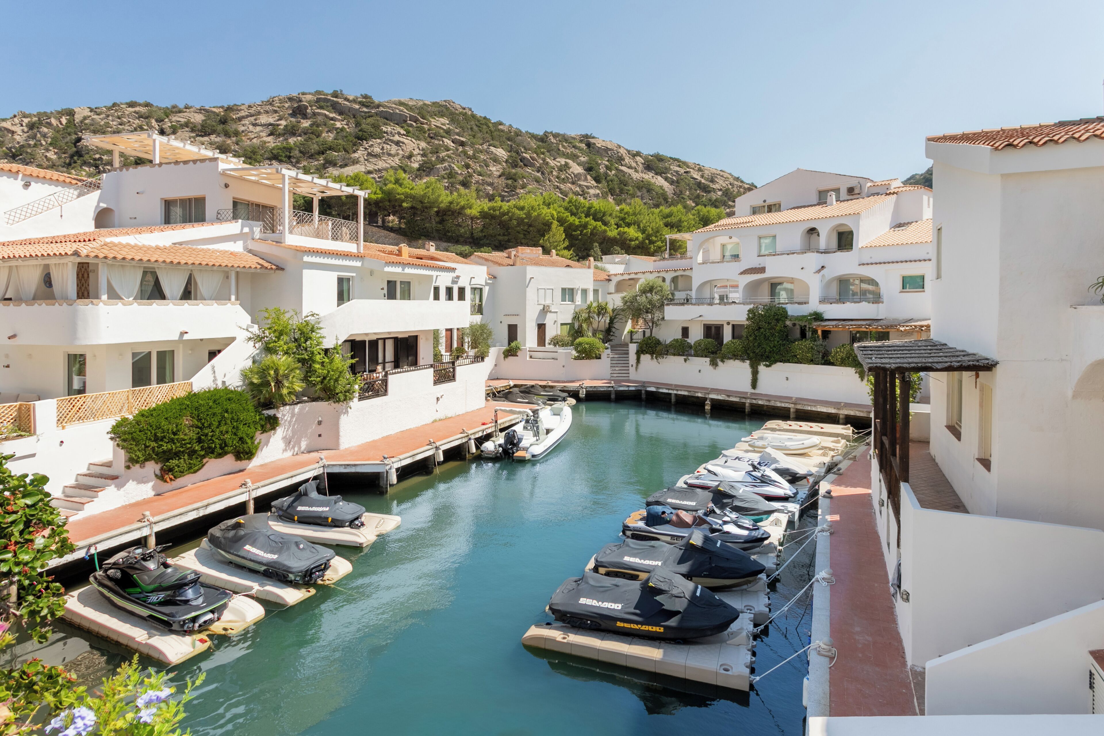 A marina with boats docked, surrounded by white buildings with red-tiled roofs, and a hilly landscape in the background.
