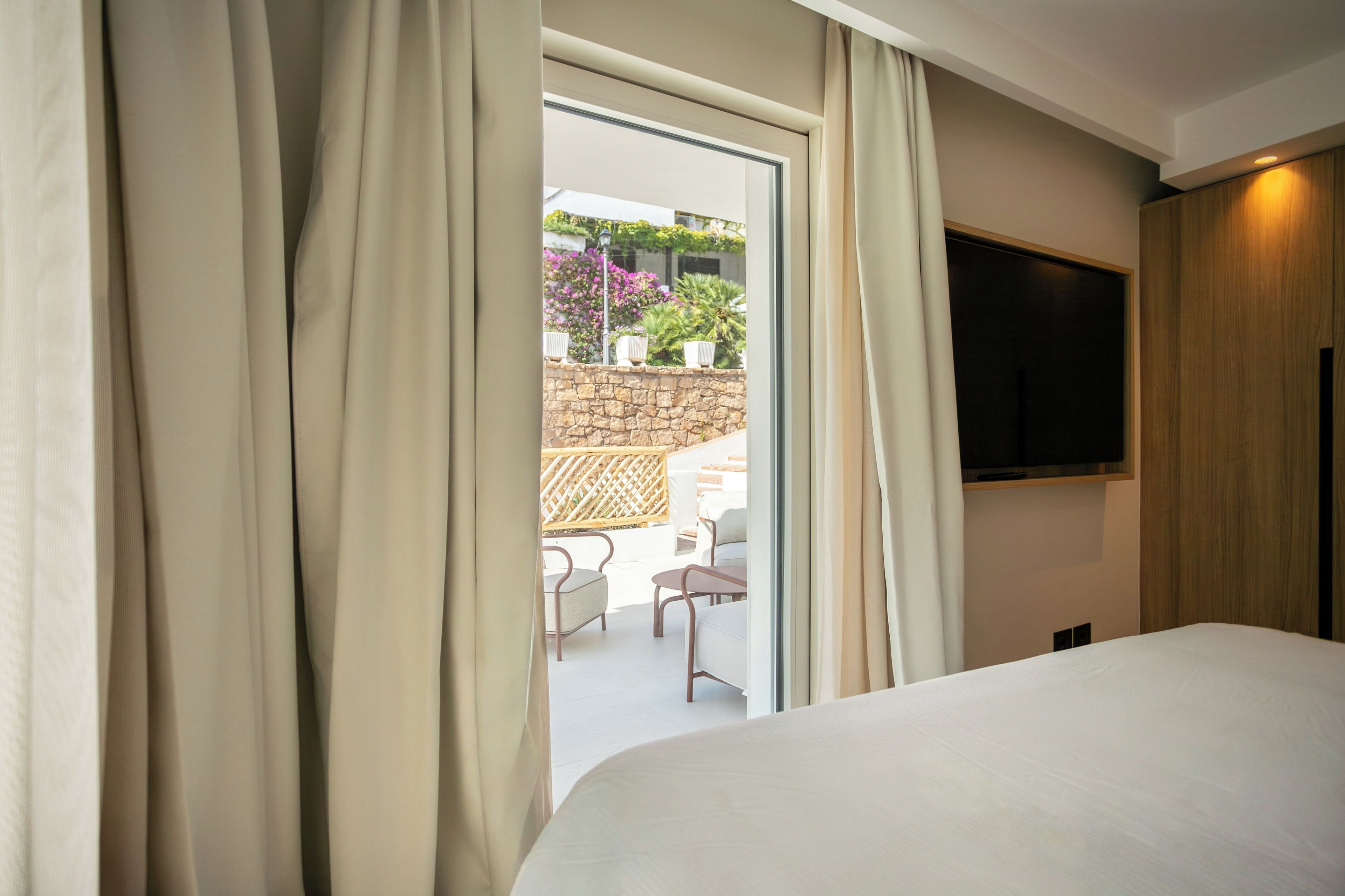 Une chambre d’hôtel avec un lit, un téléviseur fixé au mur et une vue sur un espace extérieur aménagé avec des chaises et des plantes.