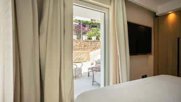 Une chambre d’hôtel avec un lit, un téléviseur fixé au mur et une vue sur un espace extérieur aménagé avec des chaises et des plantes.