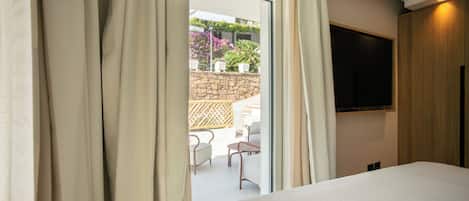 Habitación de hotel con cama, televisor montado en la pared y vista a un área exterior con sillas y plantas.