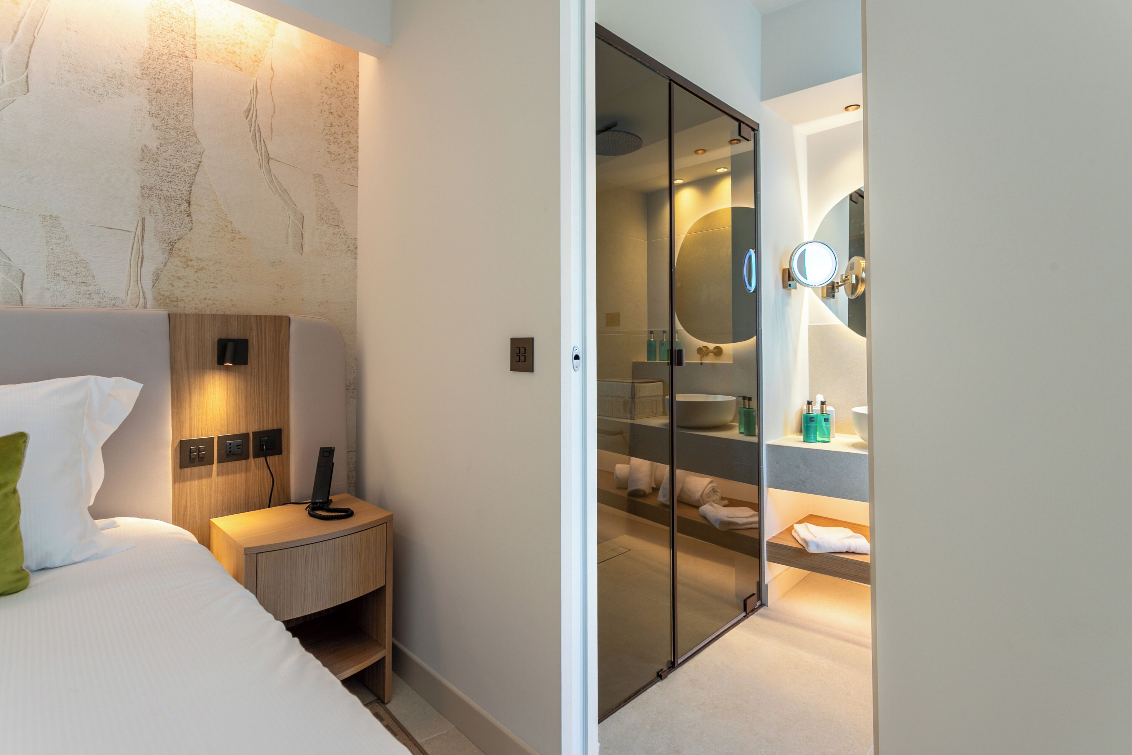 A modern hotel room with a bed, bedside table, and a glass partition leading to a bathroom with a large mirror and a sink.