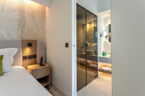 A modern hotel room with a bed, bedside table, and a glass partition leading to a bathroom with a large mirror and a sink. - Il Borgo Marina Homesuites Poltu Quatu (Arzachena)