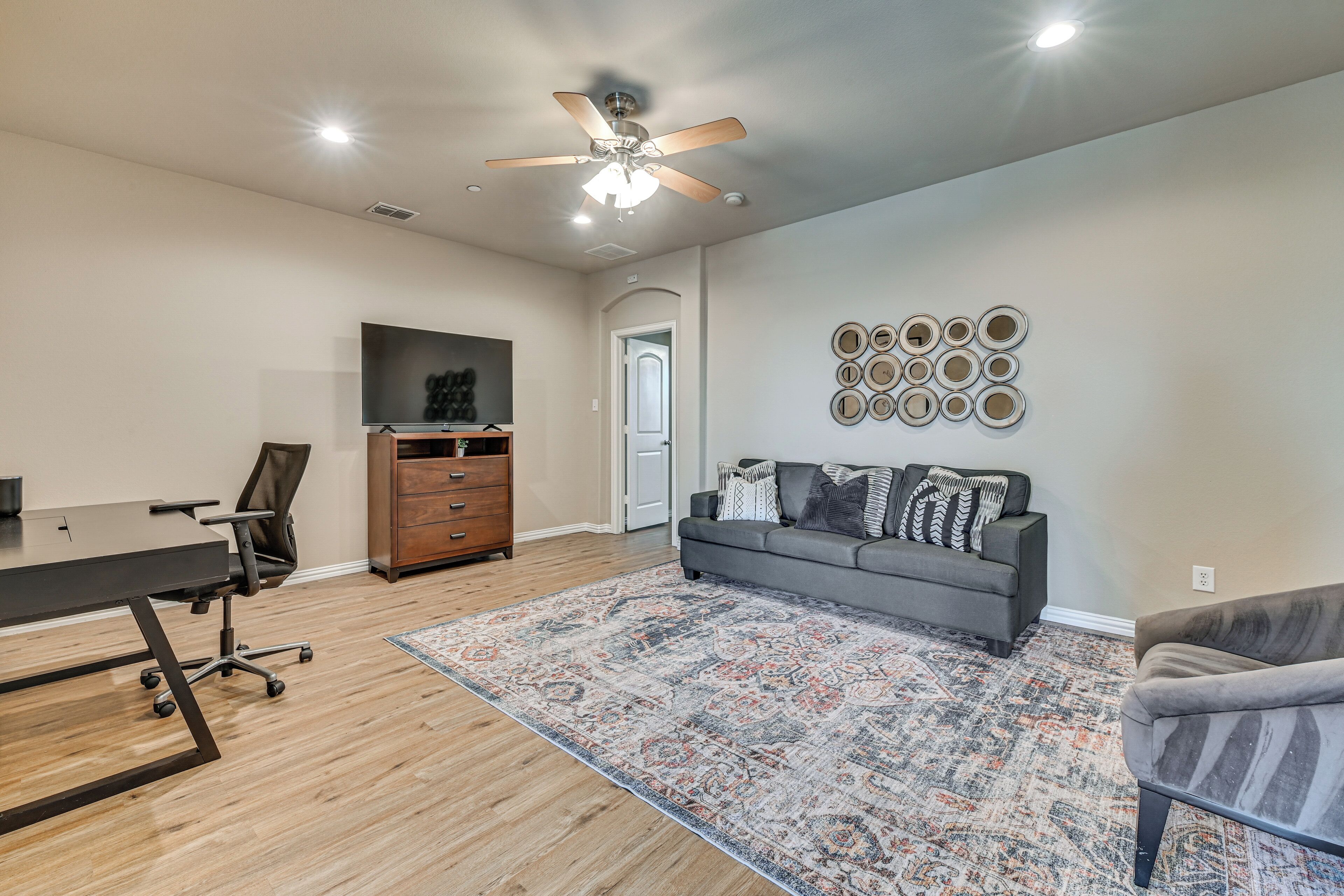 A spacious room with a ceiling fan, a large TV, a grey sofa, a patterned rug, and a wooden floor.