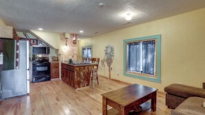A spacious living area with a wooden floor, a dining table, a sofa, and a kitchen with a microwave and stove. - Home Near Eleven Point River & Nat'l Forest! (Alton)