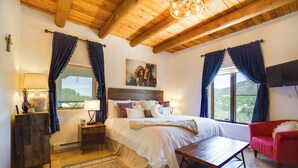 Ein Schlafzimmer mit Holzbalkendecke, einem Bett mit weißer Bettwäsche, einem roten Sessel, einem Holztisch und Blick ins Freie durch ein Fenster.