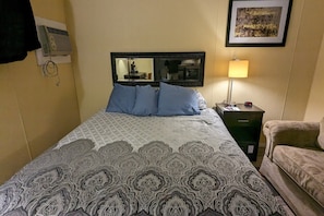 A hotel room with a bed, a nightstand, a lamp, a couch, a mirror, and a wall-mounted air conditioner.