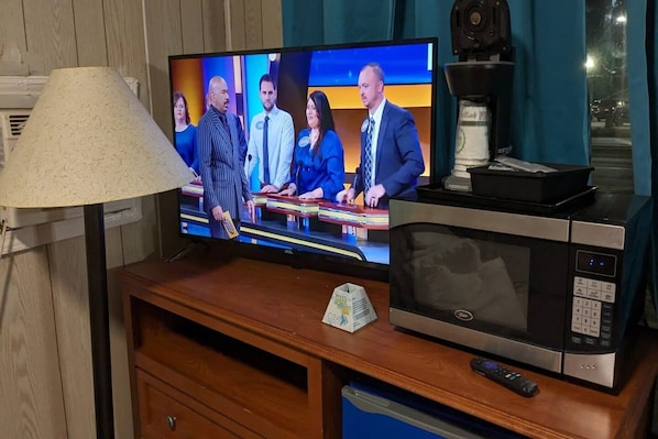 A television displaying a news program, a microwave, a remote control, and a trash bin on a wooden table.