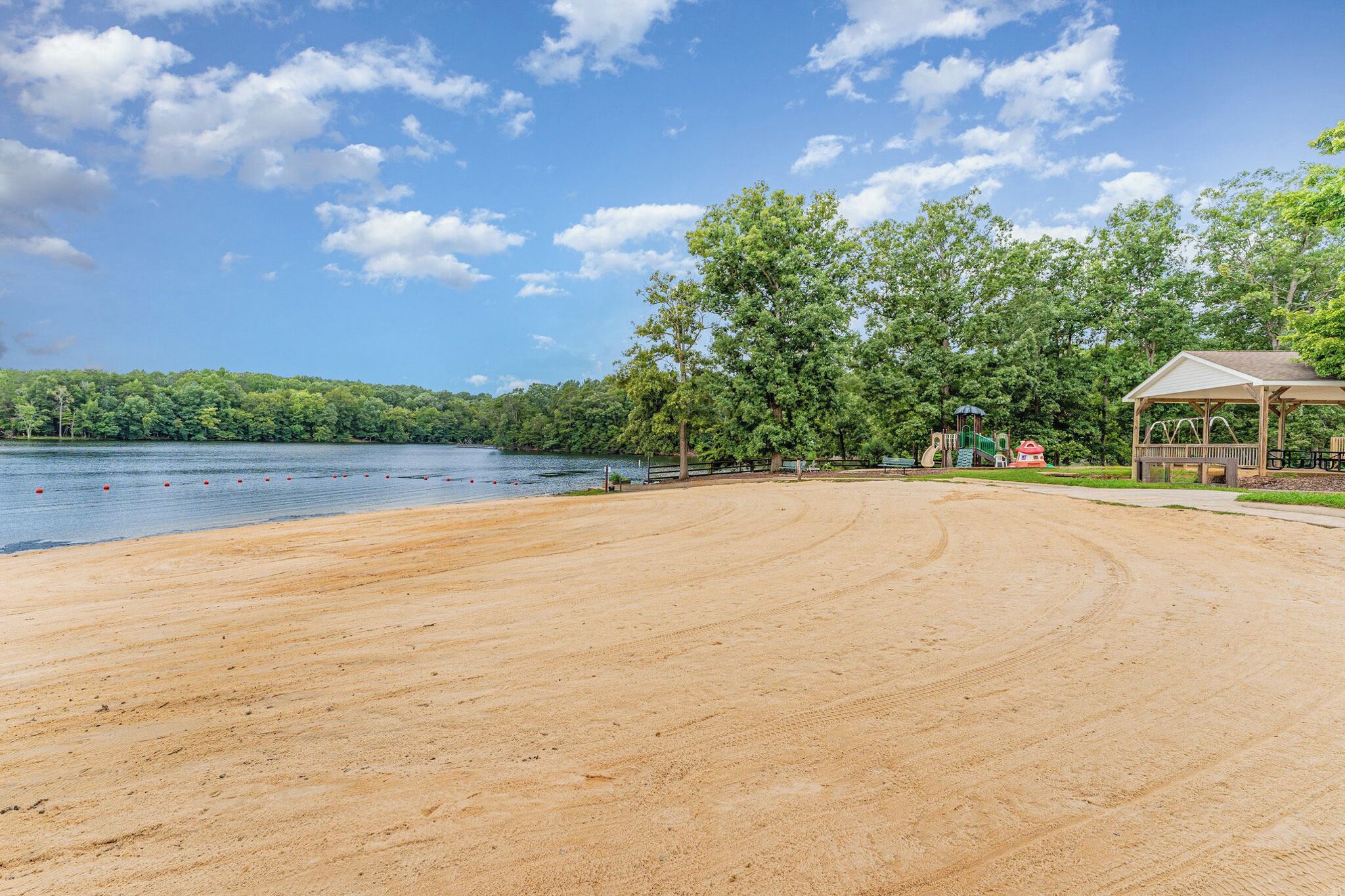 Une plage avec une zone de sable, une structure en bois et un lac bordé d’arbres en arrière-plan.