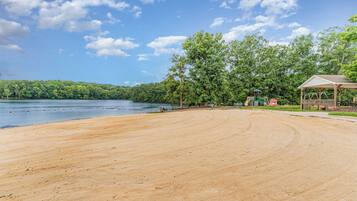 Une plage avec une zone de sable, une structure en bois et un lac bordé d’arbres en arrière-plan.