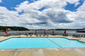 A swimming pool with lounge chairs and umbrellas, a waterfront view, and a cloudy sky.