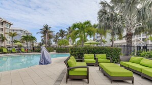 Un área junto a la piscina con sillones de descanso y sombrillas, rodeada de palmeras y un edificio.