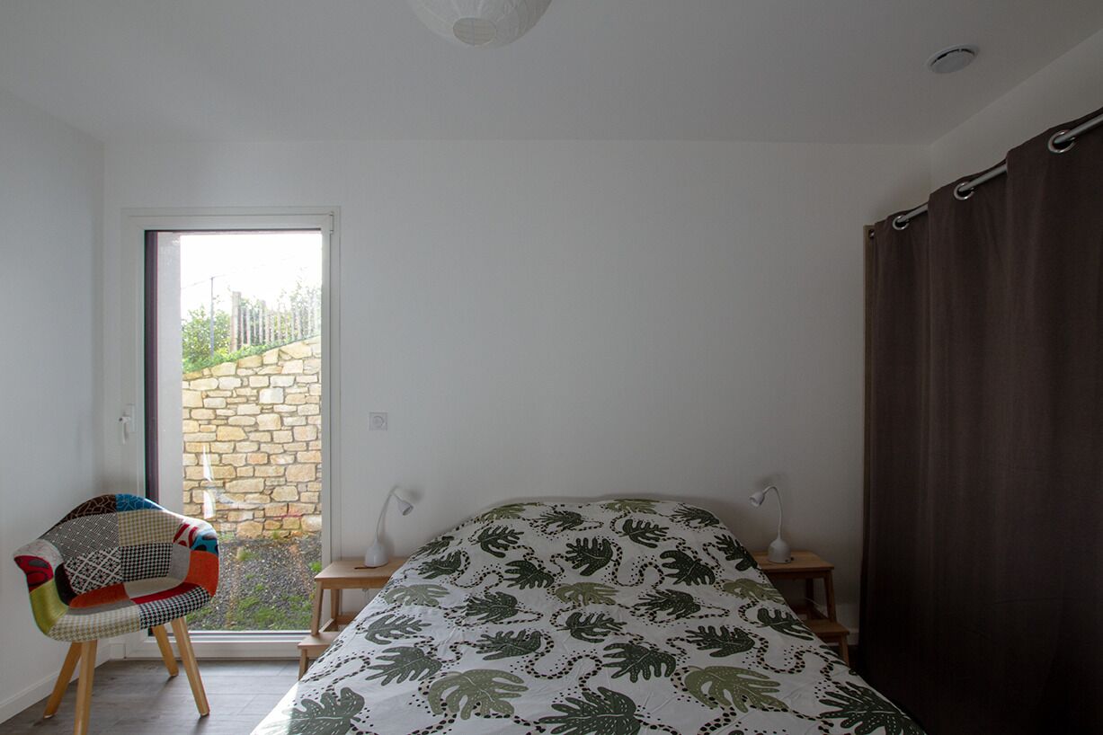Ein Schlafzimmer mit einem Bett, einem Sessel, einem Nachttisch, einer Lampe und Blick auf eine Steinsäule und Grün durch ein Fenster.