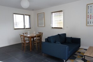 Ein Zimmer mit Esstisch, Stühlen, einer Couch und zwei Fenstern mit Jalousien.