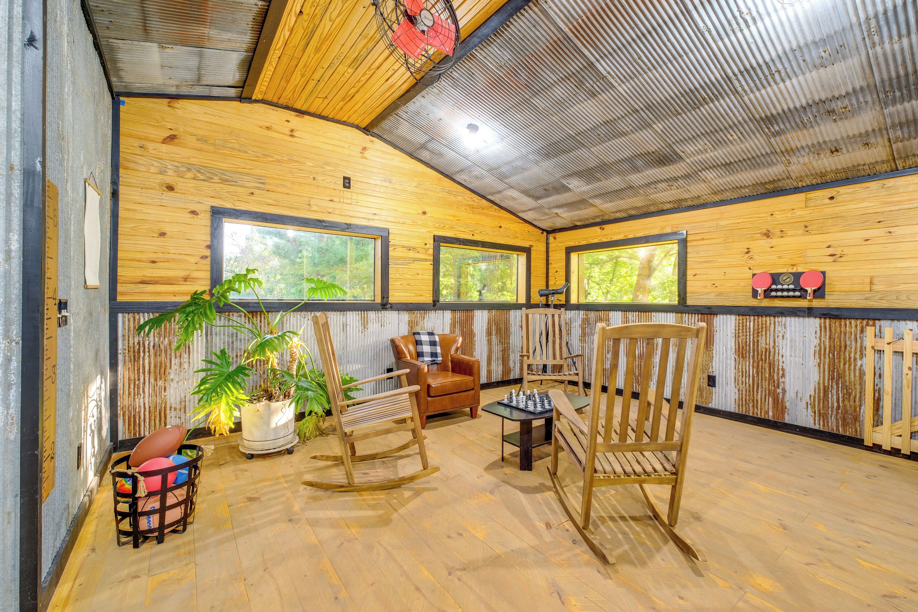 Une pièce chaleureuse avec des murs en bois, de grandes fenêtres, un fauteuil à bascule, une table d’échecs et une plante en pot.
