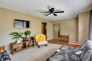 Ein Zimmer mit Deckenventilator, einem Flachbildfernseher, einem Sessel mit 'Summer'-Kissen, einem Bett mit einem Bettbezug mit Blattmuster und einer hölzernen Bank.