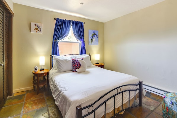 Un dormitorio con una cama, dos mesitas de noche, una ventana con cortinas azules y un cuadro colgado en la pared.