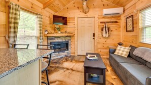 Ein Interieur einer Blockhütte mit Kamin aus Stein, Deckenventilator, montiertem Hirschkopf, einer Couch und einem Essbereich mit Stühlen.