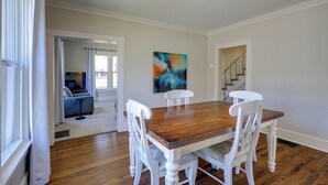 Un coin repas avec une table en bois et des chaises blanches, un tableau accroché au mur et un escalier menant à une autre pièce.