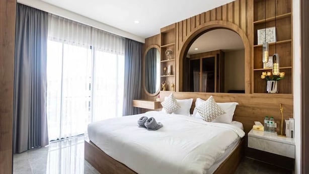 Ein modernes Hotelzimmer mit einem großen Bett, einem hölzernen Kopfteil, einer eingebauten Ablage mit Dekorationen und einem großen Fenster mit Vorhängen.