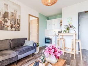 A modern living room with a sofa, a coffee table with flowers, a painting, and a kitchen area with cabinets and a dining table. - Le Sporting by Interhome (Cabourg)