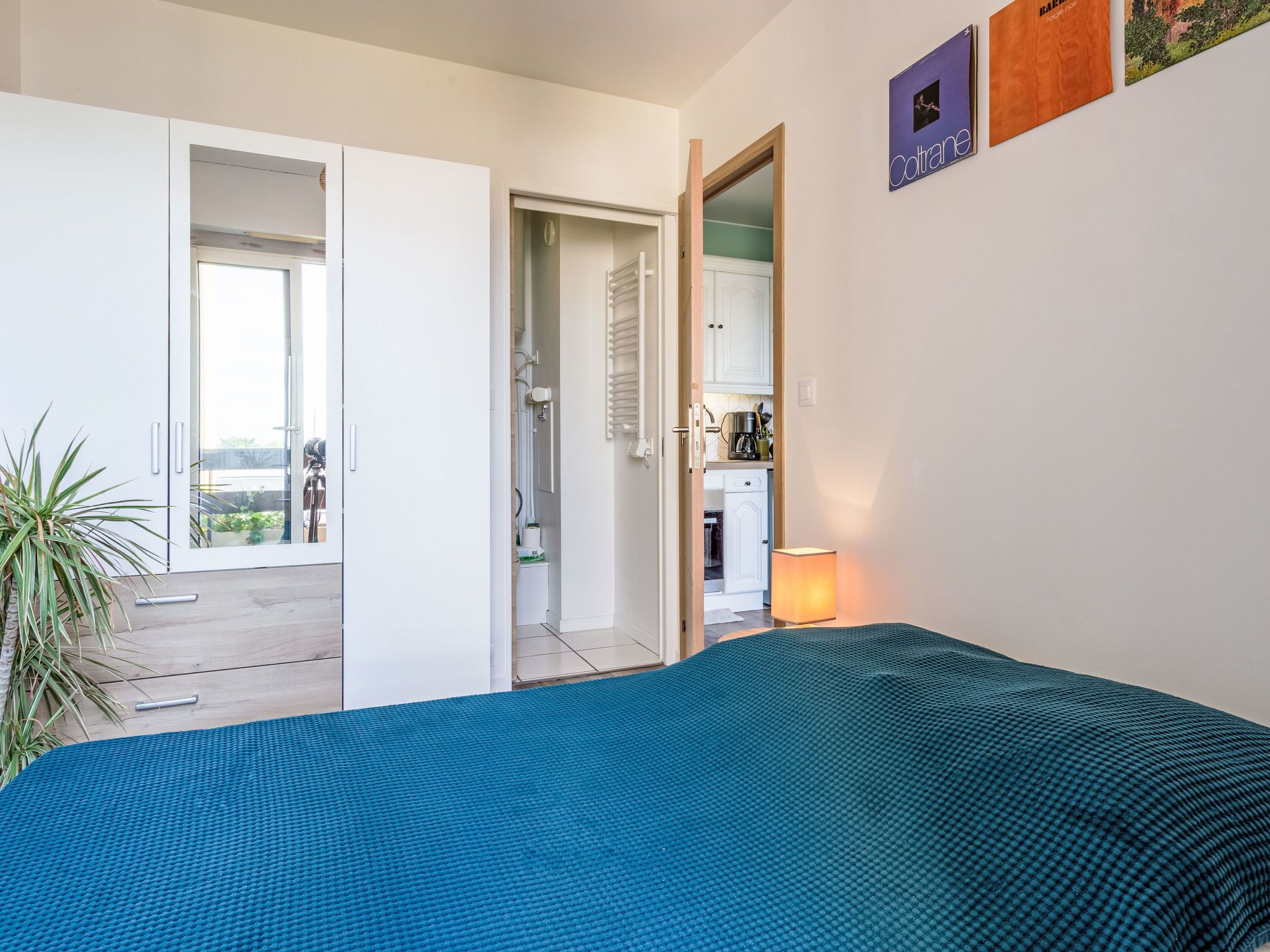 A bedroom with a blue textured bedspread, a small bedside table, a nightstand, a lamp, and a view of a balcony and kitchen area.