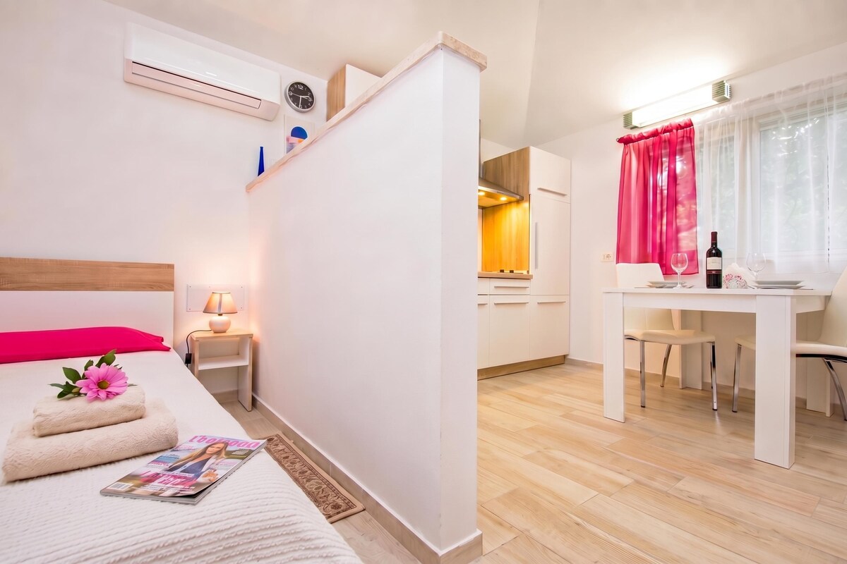A modern hotel room with a bed, bedside table, lamp, magazine, and a view of the kitchen and dining area.