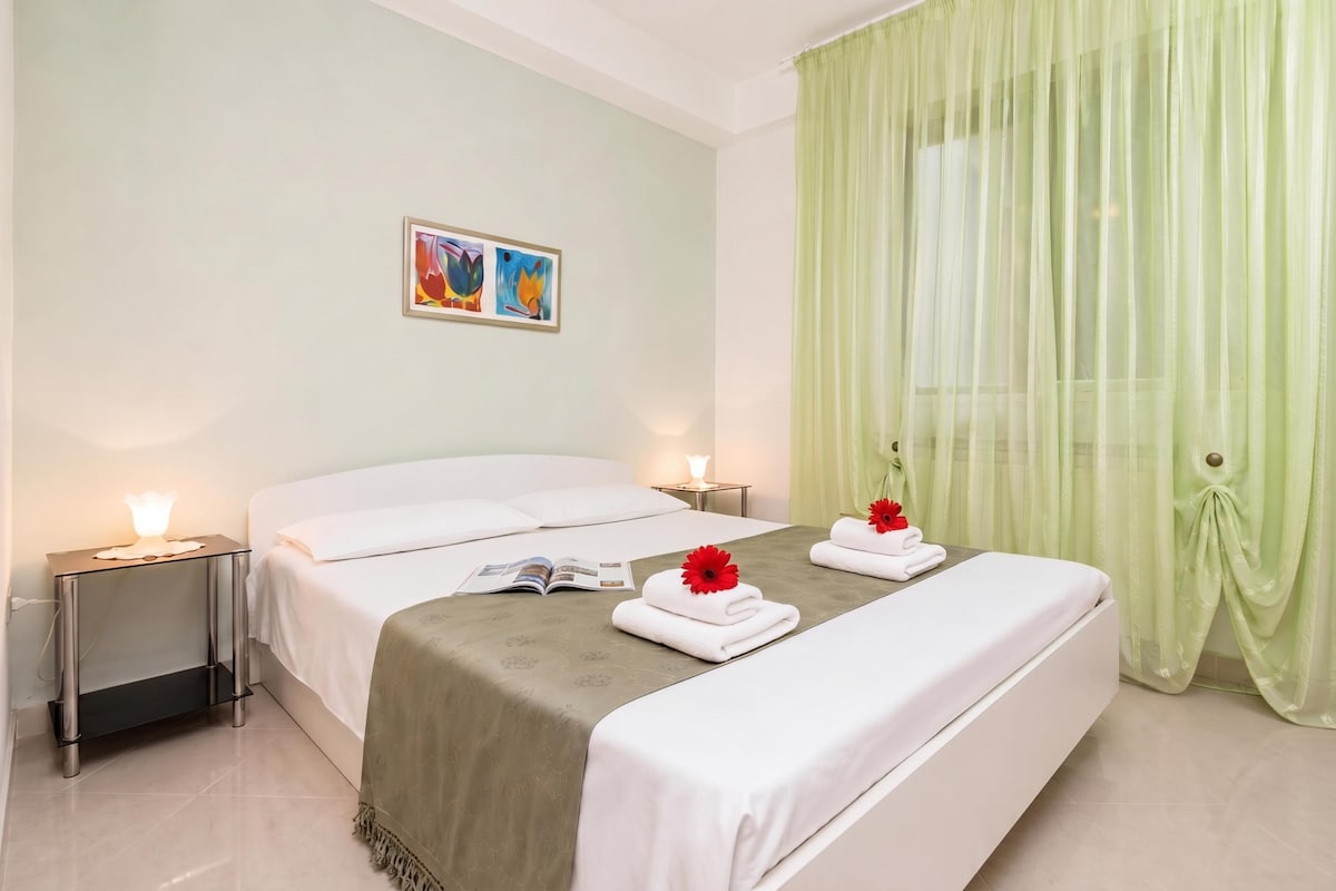 A hotel room with a bed, two bedside tables, a nightstand, a painting on the wall, and a window with sheer curtains.