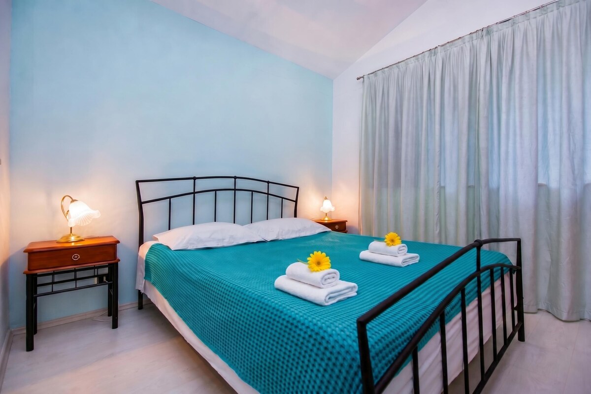 A single bed with a teal bedspread, two bedside lamps, and a window with white curtains.