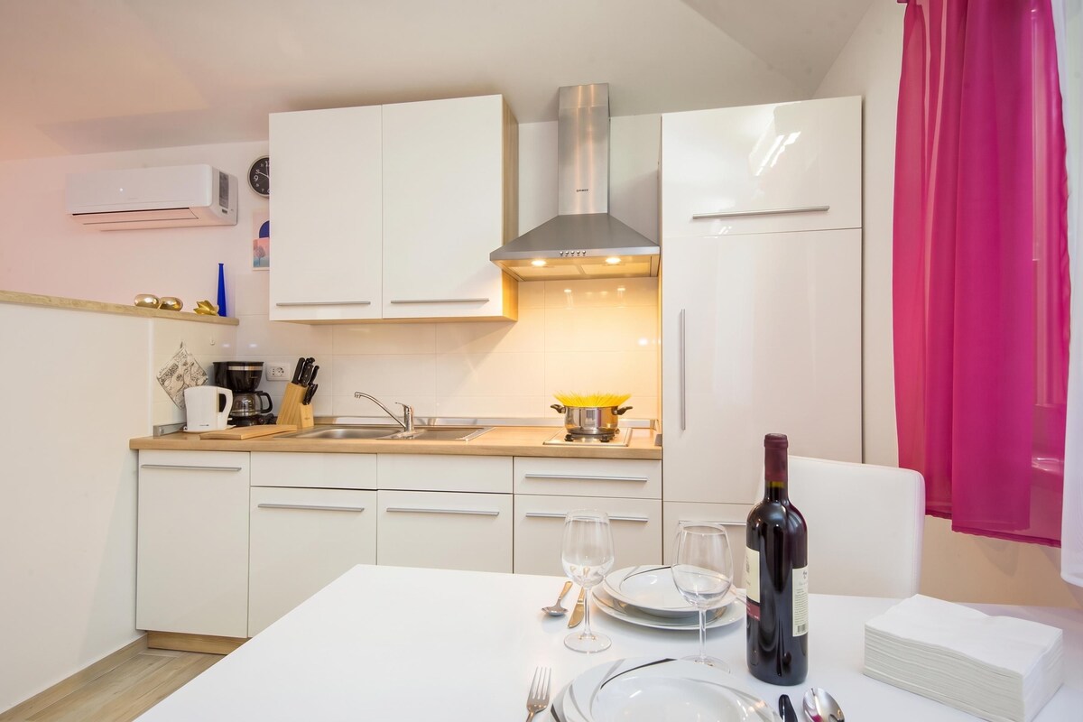 A modern kitchen with white cabinets, a stainless steel range hood, and a dining area set with wine glasses and a bottle.