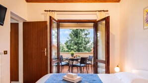 A bedroom with a bed, a bedside table, a window, and a view of an outdoor patio with a table and chairs. - Marisa Olive Grove Apartments (Labin)