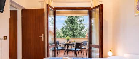 Un dormitorio con cama, mesita de noche, ventana y vistas a un patio exterior con mesa y sillas.